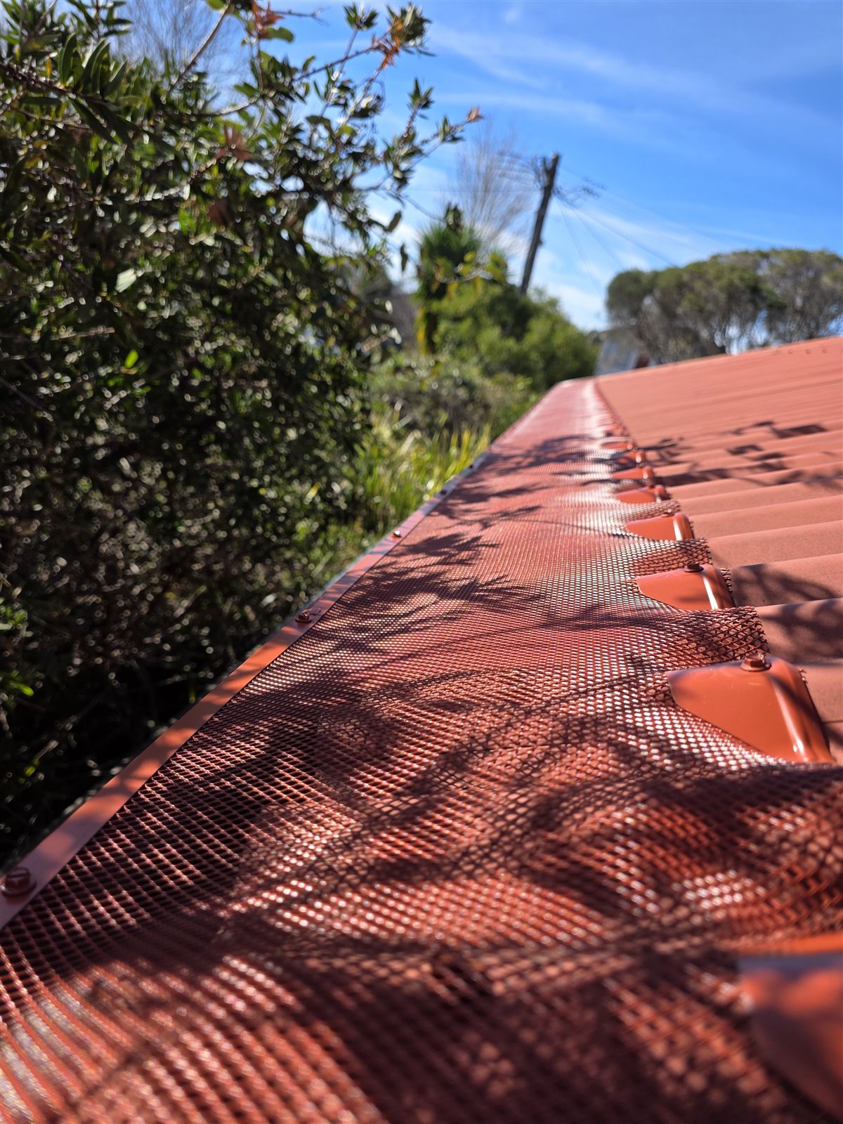 Terracotta-coloured mesh gutter guard along a tiled roof in dappled shade