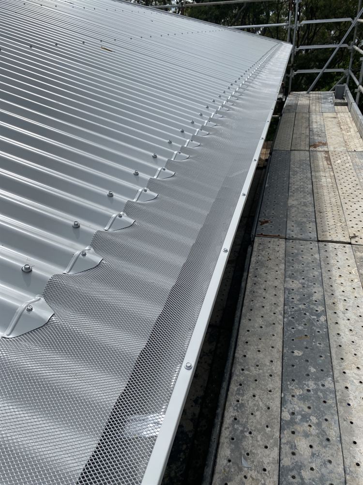 Mesh gutter guard fitted along a corrugated roof, scaffold walkway beside